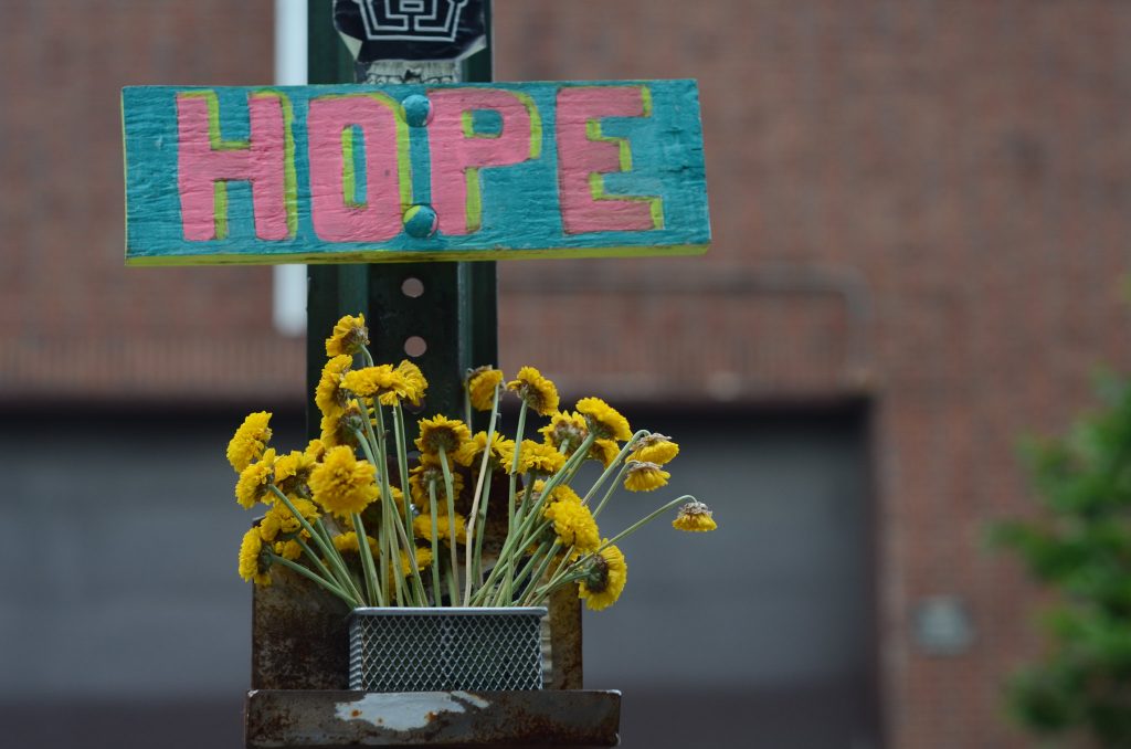 Wooden sign with "Hope" painted on it over a bouquet of yellow daisies. Hope by Gedalya AKA David Gott, used under CC 2.0 license https://creativecommons.org/licenses/by/2.0/legalcode