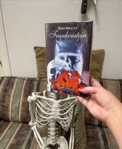 hand holding book "Frankenstein" with skull on cover in front of plastic skeleton