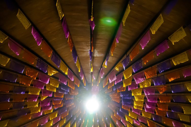 Image of ceiling with pink, yelllow and purple papel picado hung in rays from central bright light-"Danza de Papel Picado by Eneas De Troya used under cc by 2.0 license without edit or adaptation