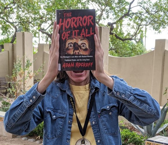 Staff holding book "The Horror of it All" in front of face